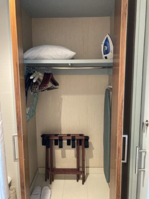 Photo of the closet area with spare pillows, iron + ironing board, and luggage rack inside. The closet doors are open.